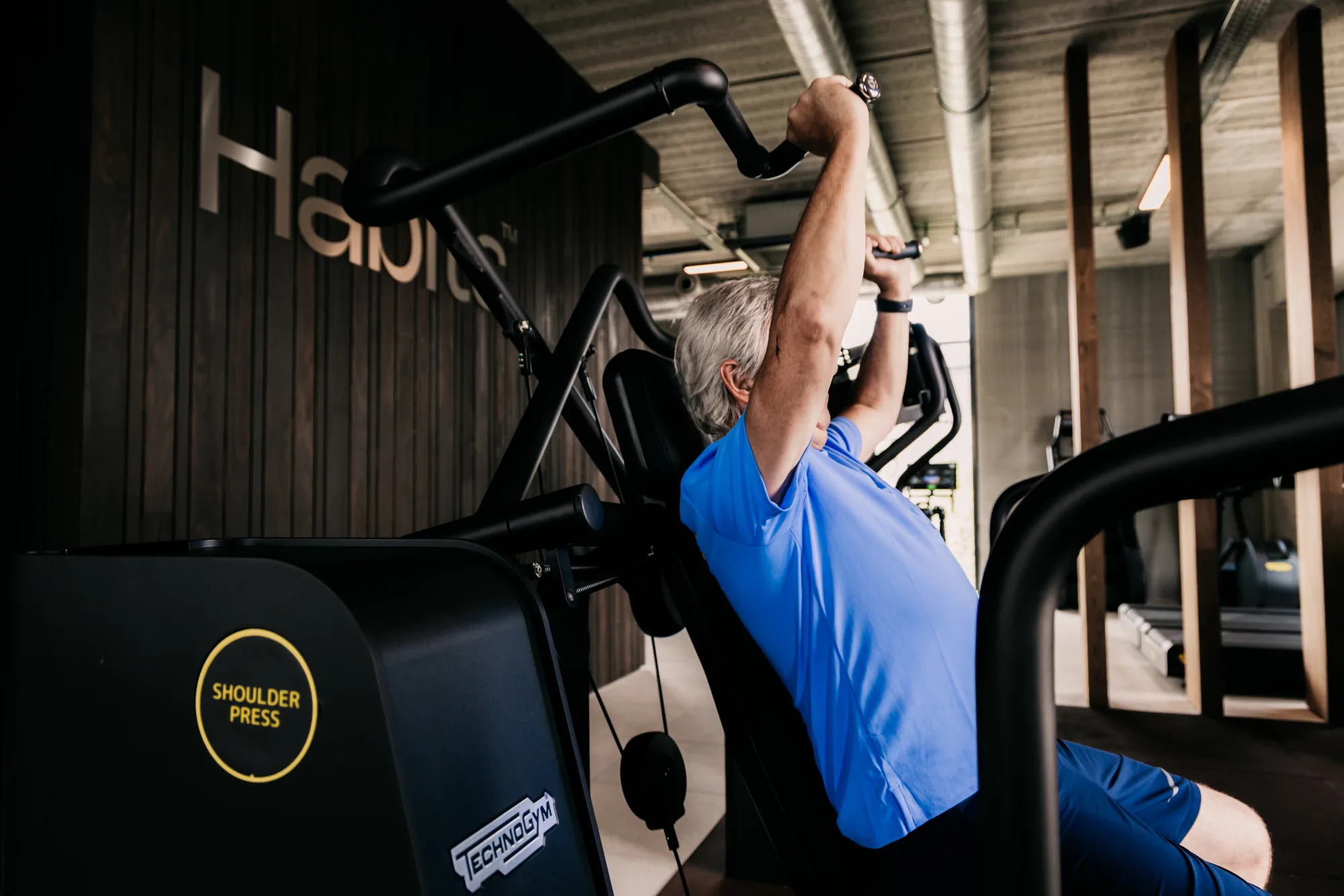 Een lid op de Technogym-shoulderpress voor de houten Habits-wand